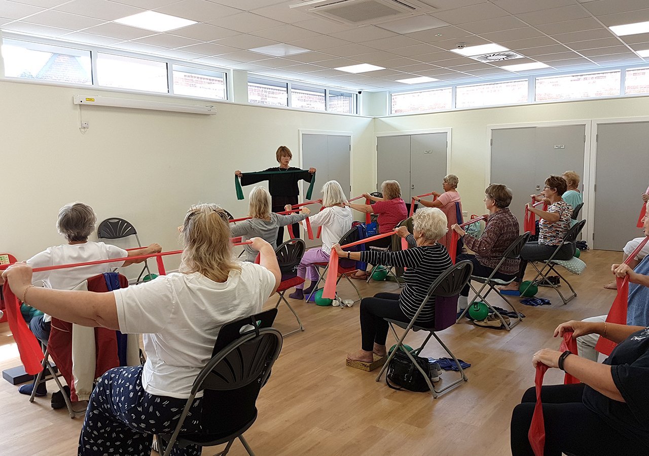 Wide view of one of Liz's classes in a bright community hall: participants seated on chairs stretching red resistance bands, Liz at the far end facing them