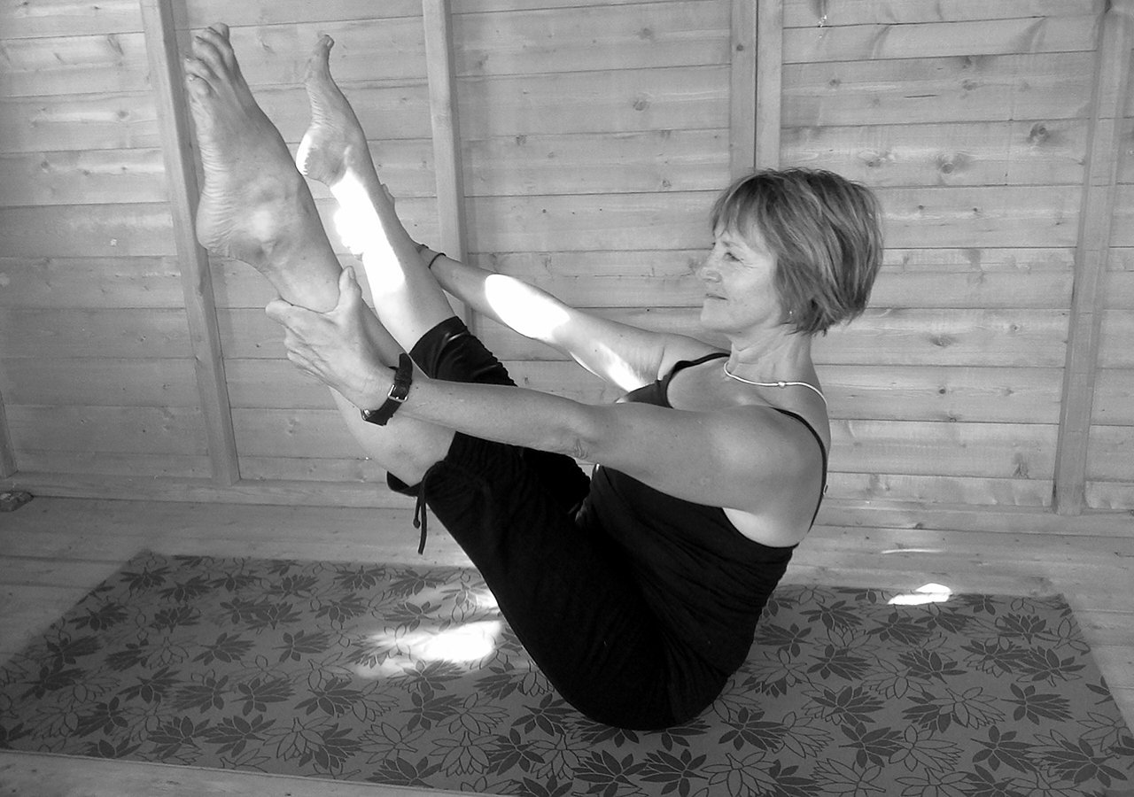 Liz Downes smiling in a full boat pose on a patterned mat in a timber-clad studio, sunlight across the wall