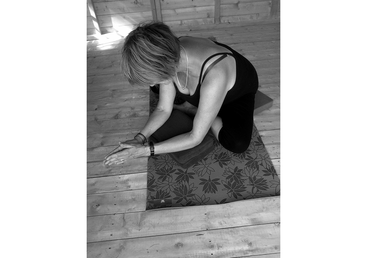 Liz seated on a mat in a twist, one arm extended along the floor, timber wall behind
