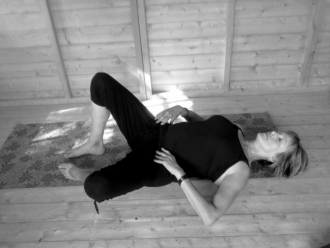 Liz lying on her side on a mat, top leg bent and held in a supported side-lying stretch