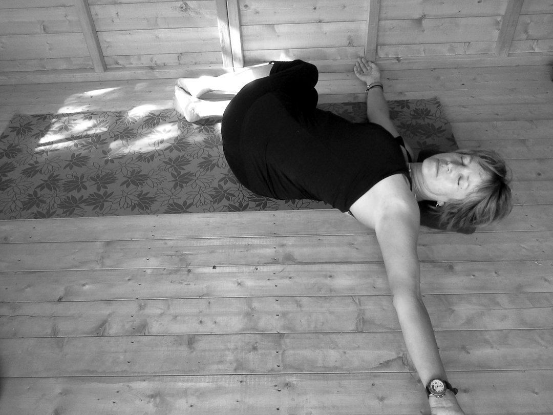 Liz lying on the wooden floor in a supine twist, one arm out, head turned toward the camera